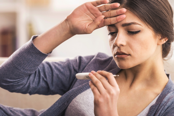 Young lady on couch with hand on her forehead while looking at a thermometor