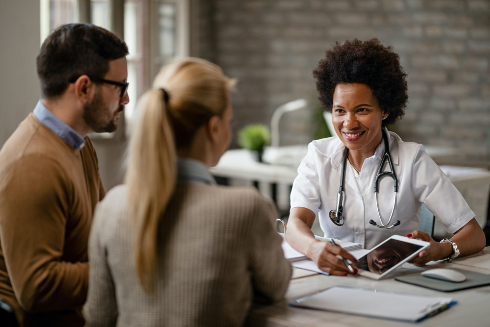 Happy black female doctor talking to a couple and showing them medical test results on a touchpad during consultations