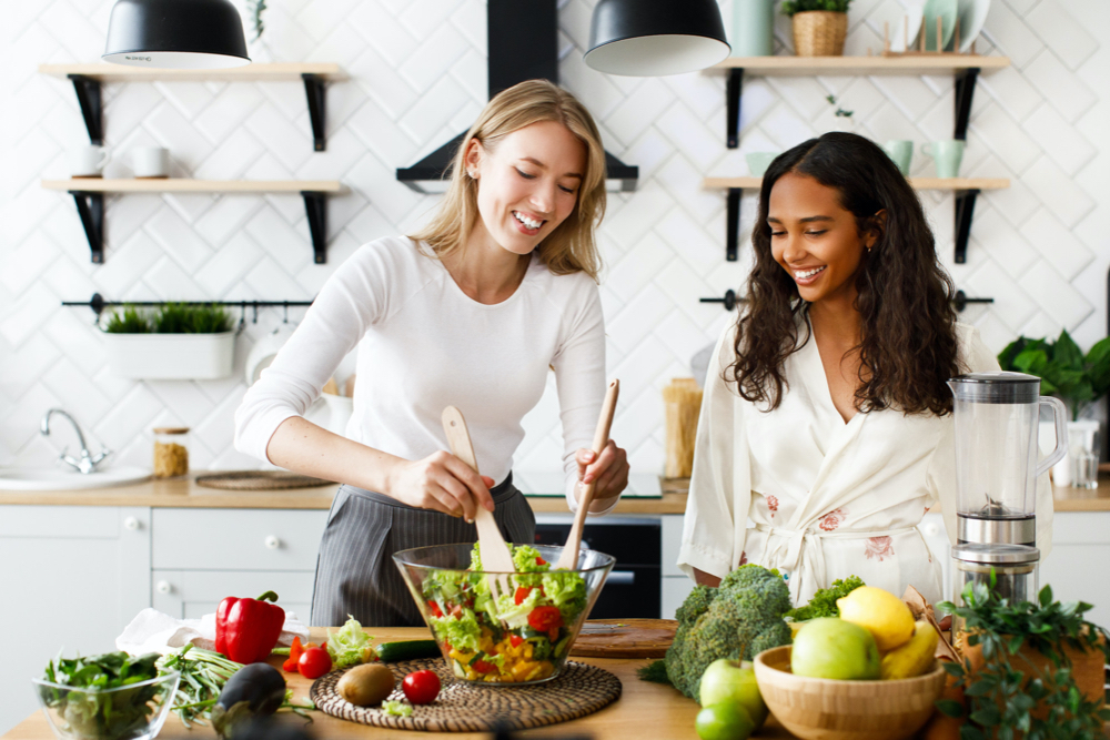 photo two women of different nationalities are smiling and cooking a salad in the kitchen eating better to lower their apob naturally