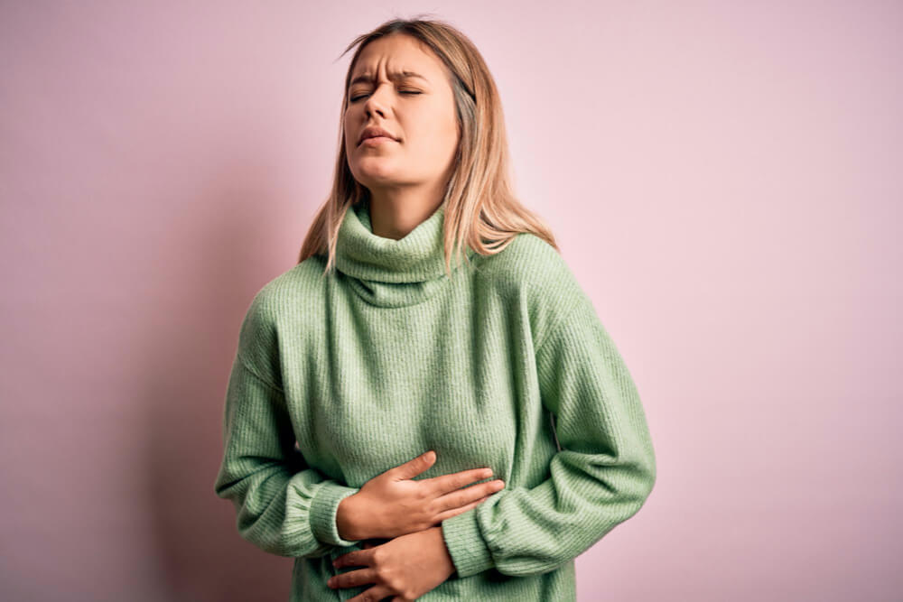 Young beautiful blonde woman wearing winter wool sweater over pink isolated background with hand on stomach because nausea painful disease feeling unwell Ache concept
