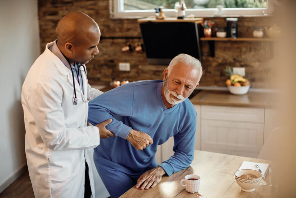 Black doctor assisting senior man to get up from the chair at nursing home
