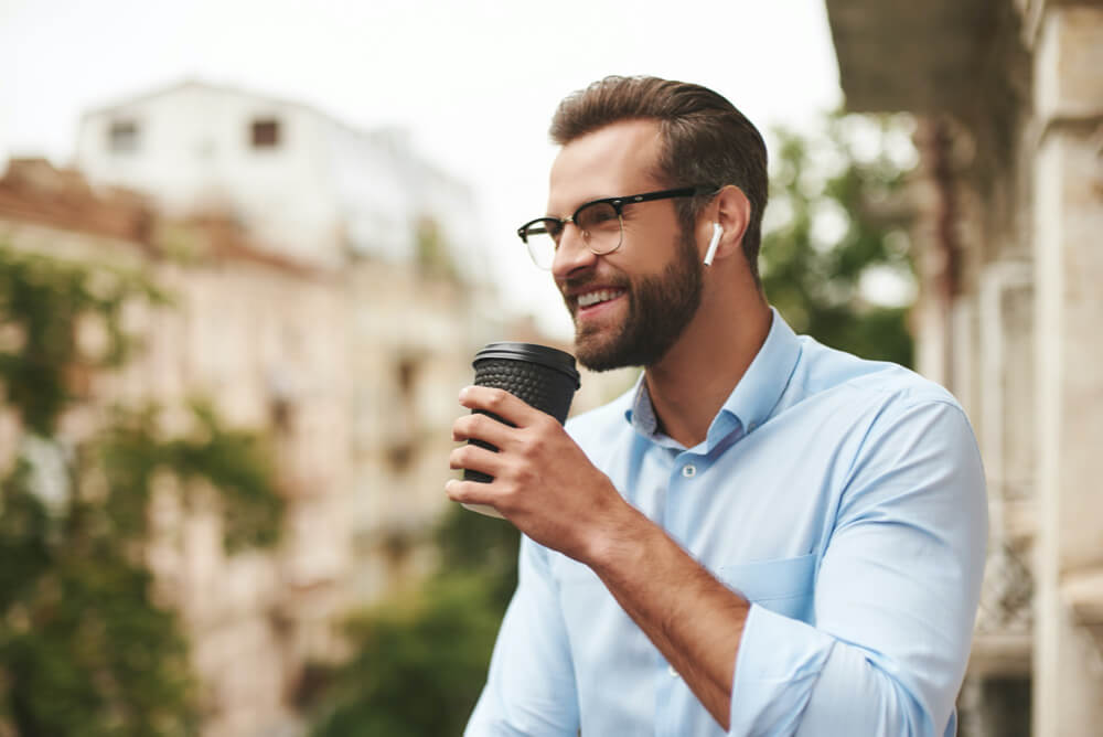 Young and handsome man in eyeglasses and headphones holding cup of coffee and talking with friend